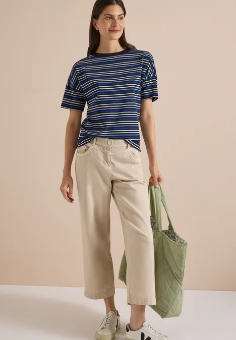 Kobieta w niebiesko-białej koszulce w paski, beżowych culottes i zielonej torbie