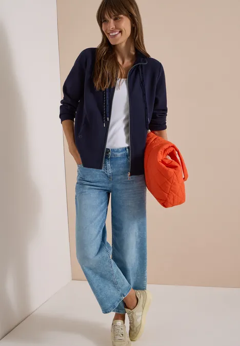 Langarm Jacke mit Ottoman Struktur urban dark blue Eine lächelnde Frau in Jeans, weißem Shirt, blauer Jacke und Turnschuhen hält einen orangen Pullover.