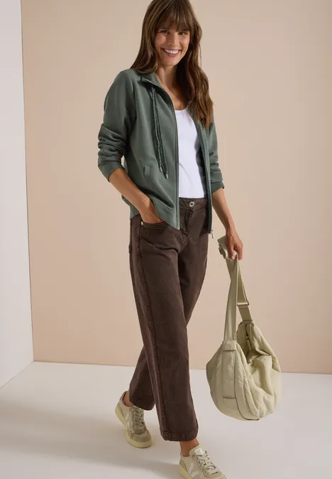 Langarm Jacke mit Ottoman Struktur dried olive green Junge Frau lächelt, trägt weiße Bluse, grüne Jacke, braune Hose, beigetöne Tasche und Sneaker.