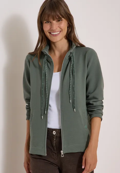 Langarm Jacke mit Ottoman Struktur dried olive green Lächelnde Frau in grüner Kapuzenjacke und weißem Top vor hellem Hintergrund.