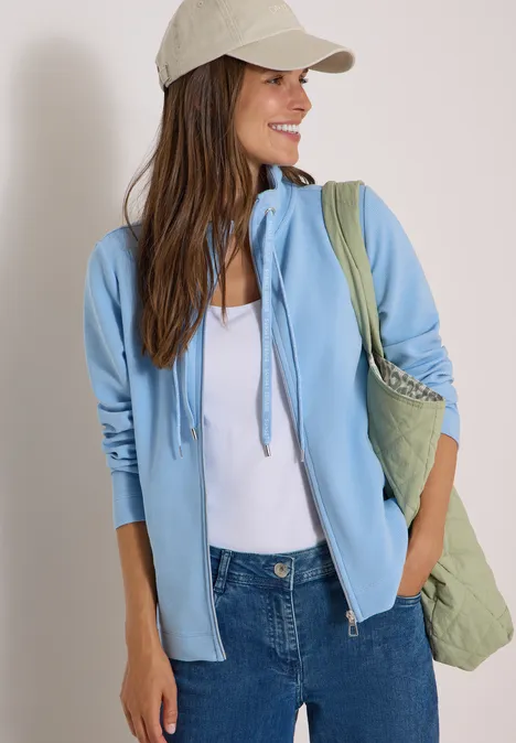 Jas met lange mouwen en ottomanstructuur easy blue Een lachende vrouw met een beige pet, lichtblauwe hoodie, wit topje, spijkerbroek en een groene tas.