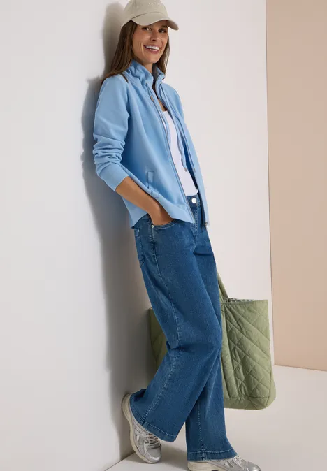 Jas met lange mouwen en ottomanstructuur easy blue Vrouw leunt lachend tegen een muur, draagt lichtblauw overhemd, wit T-shirt, wijde jeans, witte sneakers, pet en groene tas.
