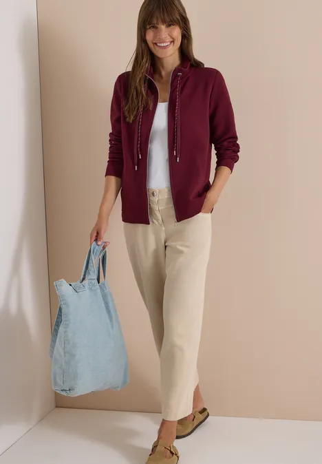 Frau lächelt, trägt eine weinrote Strickjacke, graues Shirt, helle Hose und hält eine hellblaue Stofftasche.