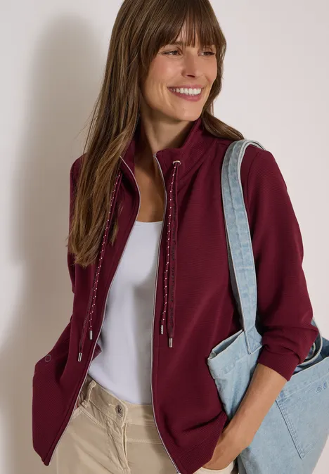 Frau lächelt, trägt weinrote Sweatjacke, weißen Top, beige Hose und hellblauen Shopper.