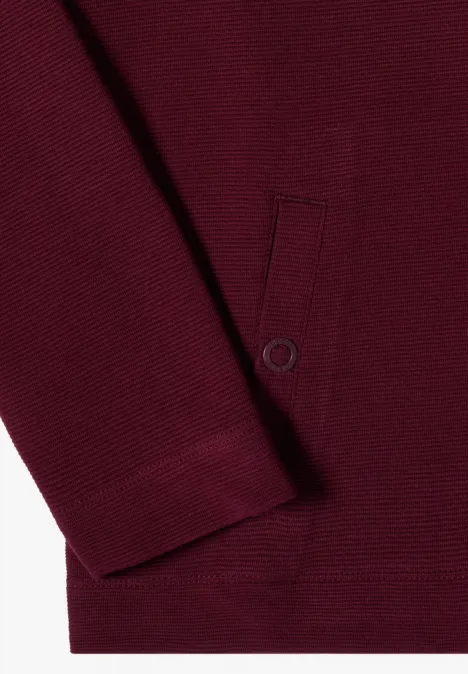 Nahaufnahme eines bordeauxroten Pullovers mit Rippbündchen und aufgesetzter Tasche mit Druckknopf.