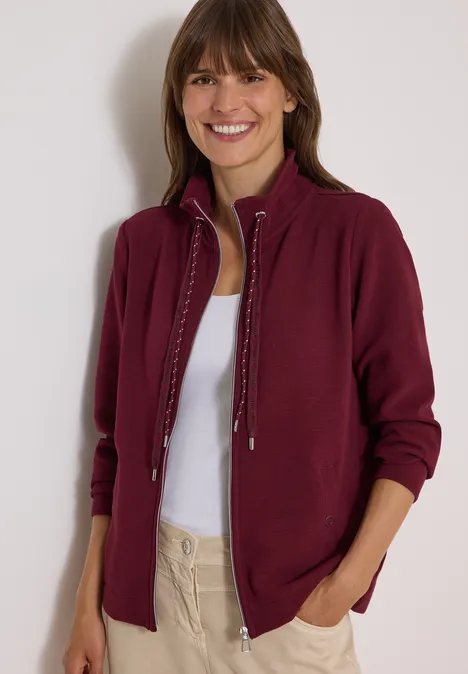 Frau in weinrotem Cardigan, weißem Top und beigen Hosen lächelt mit Händen in den Taschen vor hellem Hintergrund.