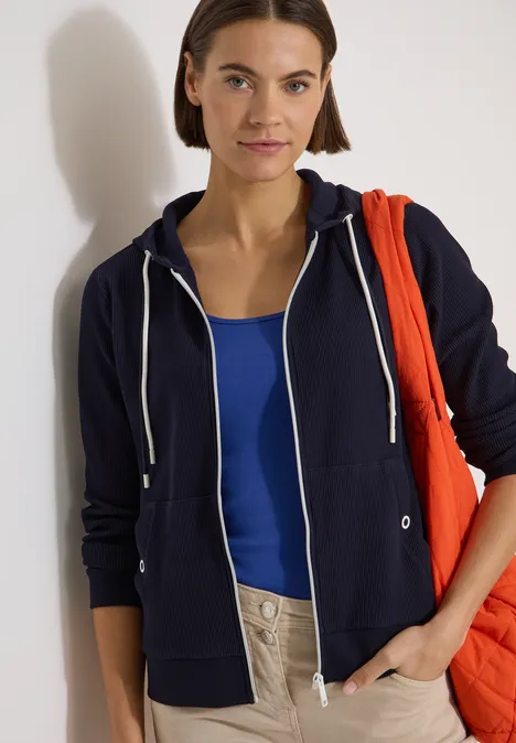 Langarm Jacke mit Kapuze und Struktur urban dark blue Person trägt dunkelblauen Hoodie, blaues Top und helle Hose, hält rote Tasche über der Schulter.