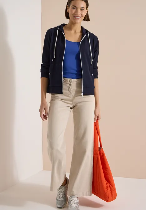 Langarm Jacke mit Kapuze und Struktur urban dark blue Frau trägt beige weite Hose, blaues Top, dunkelblaue Kapuzenjacke, graue Sneaker und orange Tasche.