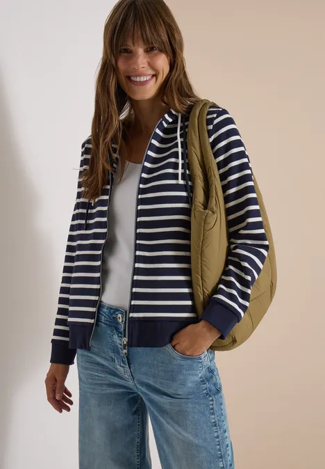 Junge Frau trägt gestreiften Kapuzenpullover, weiße Bluse, Jeans und olivgrüne Tasche, sie lächelt.