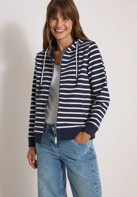 Lächelnde Frau in gestreifter Kapuzenjacke, weißem Top und Jeans, steht mit Händen in den Taschen.