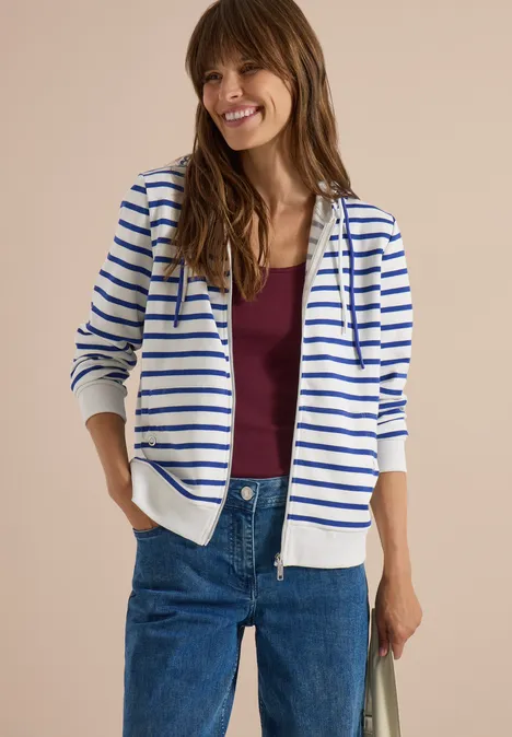 Frau in weißem Hoodie mit blauen Streifen, weinrotem Shirt und Jeans, lächelnd