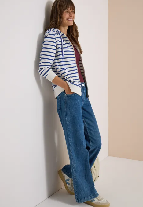 Frau lehnt lässig an einer Wand, trägt gestreiften Cardigan und weite Jeans, lächelt.