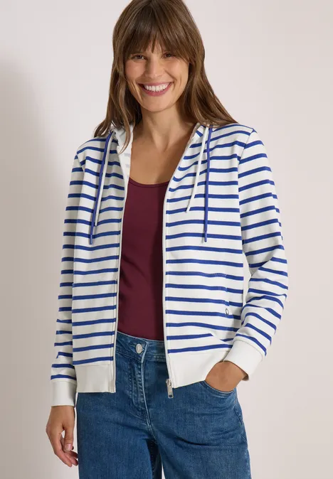 Fröhliche Frau im weißen Kapuzenpullover mit blauen Streifen, rotem Top und Jeans.