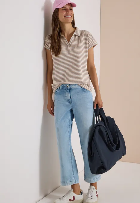 Kurzarm Poloshirt mit Streifenstruktur creamy mocha Frau lehnt an Wand, trägt gestreiftes Top, weite Jeans, weiße Sneakers, rosa Cap und hält große blaue Tasche.