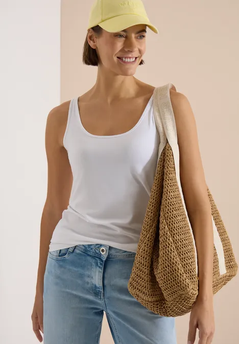 Haut long à col rond, de couleur unie White Frau in weissem Top und Jeans trägt eine gelbe Kappe und eine große gehäkelte Tasche, sie lächelt.
