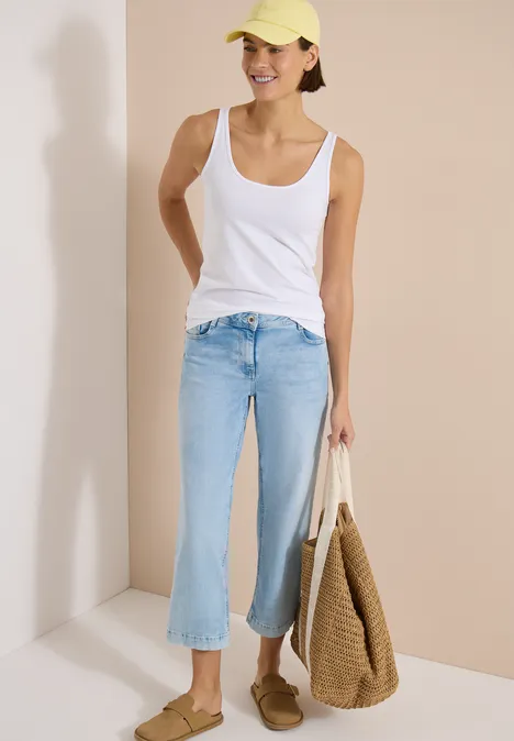Haut long à col rond, de couleur unie White Frau in weißem Tanktop, hellblauen Culotte-Jeans, Strohtasche und Mokassins steht vor hellem Hintergrund.