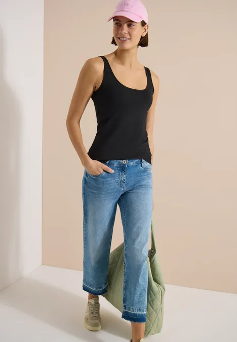 Frau in schwarzem Tanktop, weiten blauen Jeans, beigen Stiefeln und grüner Tasche.