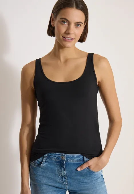 Frau in schwarzem Tanktop und blauen Jeans, eine Hand in der Hosentasche, neutraler Hintergrund.