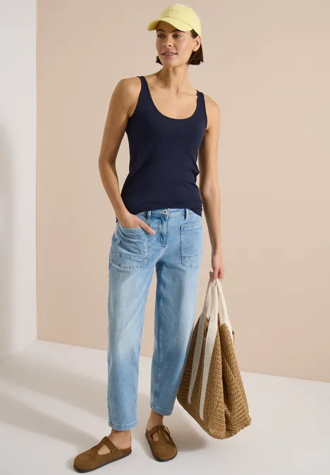 Frau in marineblauem Top und hellblauen Jeans trägt eine große gewebte Tasche und braune Sandalen.