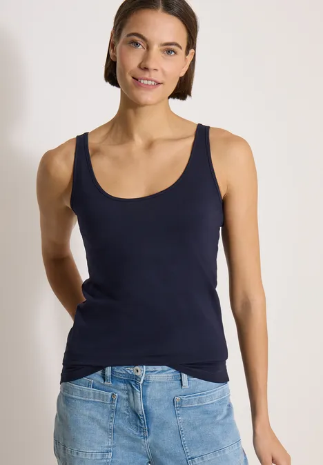 Frau in marineblauem Tanktop und Jeans, stehend vor weißem Hintergrund, lächelnd, Hände am Rücken.