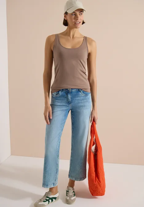 Haut long à col rond, de couleur unie cocoa taupe Eine Person in braunem Top, blauen Jeans und Sneakers hält eine rote Tasche vor hellem Hintergrund.