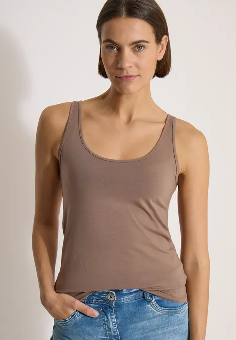 Haut long à col rond, de couleur unie cocoa taupe Frau mit braunem Tanktop und Jeans, steht vor weißem Hintergrund, eine Hand in der Hosentasche.