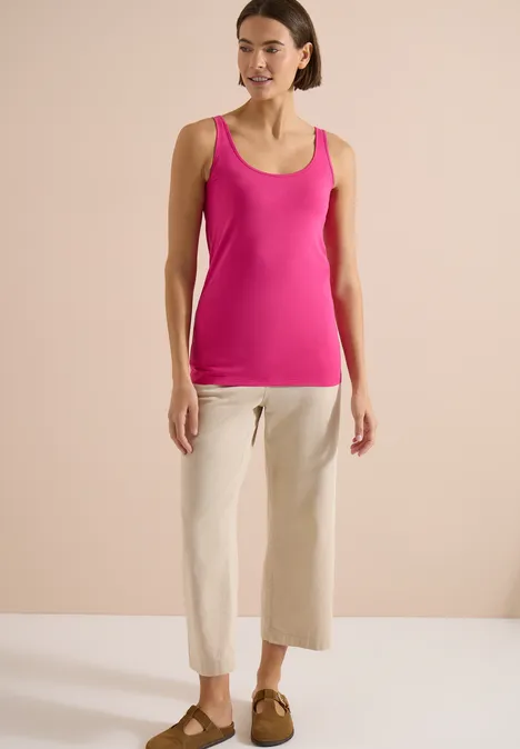 Haut long à col rond, de couleur unie pink flash Frau in pinkem Tanktop und beigen, weiten 7/8-Hosen vor neutralem Hintergrund.