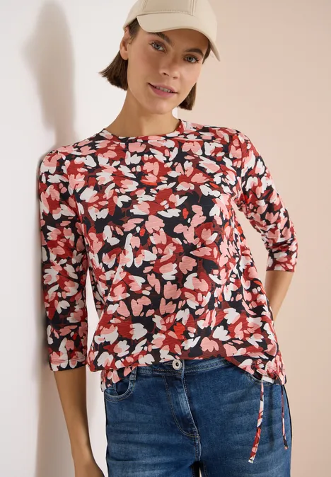 T-shirt à manches 3/4, col rond et imprimé russet red Frau in Blütenmuster-Bluse mit 3/4-Ärmeln und Jeans vor hellem Hintergrund.