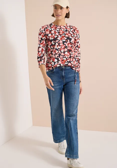 T-shirt à manches 3/4, col rond et imprimé russet red Frau trägt weite blaue Jeans, rote geblümte Bluse und weiße Sneaker vor neutralem Hintergrund.