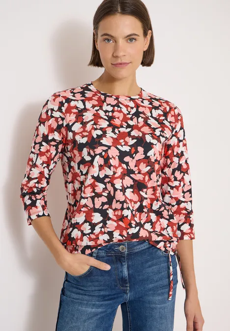 T-shirt à manches 3/4, col rond et imprimé russet red Frau trägt roten, schwarz-weiß Blumenpullover mit 3/4-Ärmeln und Bindeband, kombiniert mit blauen Jeans.