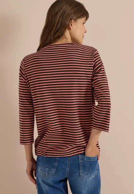 Person von hinten in rostrotem 3/4-Arm-Shirt mit schwarzen Streifen und blauer Jeans vor beigem Hintergrund.