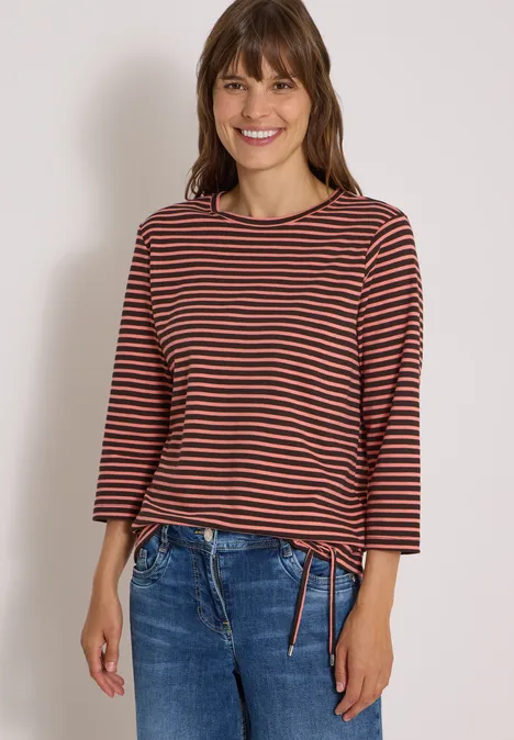 Junge Frau in gestreiftem Langarmshirt und Jeans lächelt vor neutralem Hintergrund.
