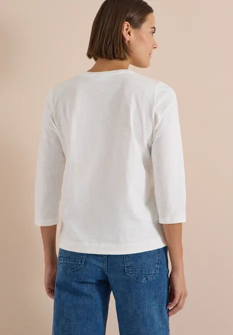 Frau von hinten in weißem 3/4-Arm-Shirt und Jeans vor beigem Hintergrund.