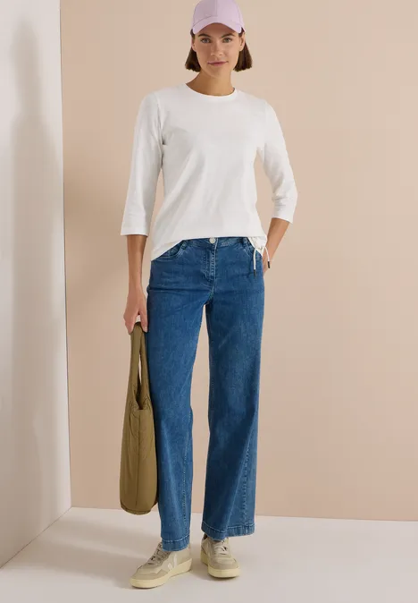 Frau mit weißem 3/4-Shirt, blauen Jeans, beigen Sneakern und olivgrünem Beutel vor beiger Wand