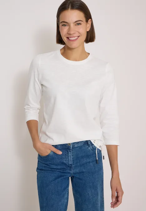 Junge Frau in weißem Langarmshirt und blauen Jeans steht lächelnd vor neutralem Hintergrund