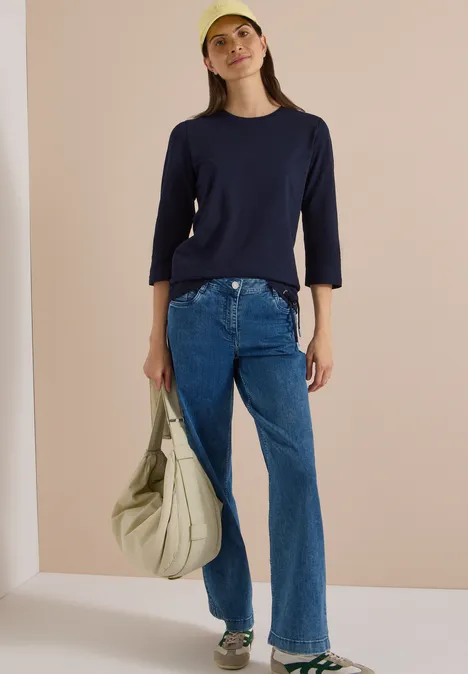 Frau in dunkelblauem Shirt, weiten Jeans und gelber Cap hält eine große, beige Tasche vor hellem Hintergrund.