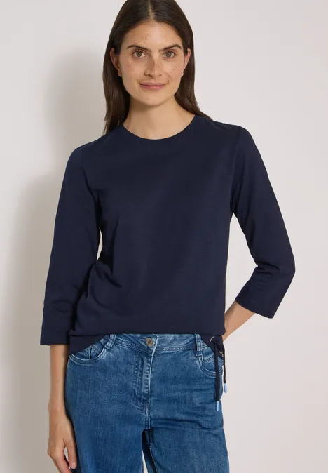 Frau trägt dunkelblaues Langarmshirt mit Seitenschnürung und blaue Jeans.