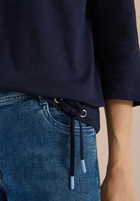 Ausschnitt zeigt dunkelblauen Sweatshirt-Saum mit Kordelzug und hellblaue Jeans, Hand am Hüftbereich.