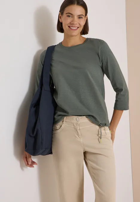 Lächelnde Frau in grünem 3/4-Arm-Shirt, beiger Hose und blauer Umhängetasche.