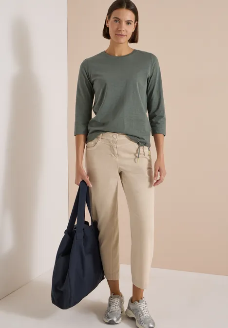 Frau in grauem Dreiviertelarm-Shirt, beige Hose und Sneakers hält eine große dunkelblaue Tasche.