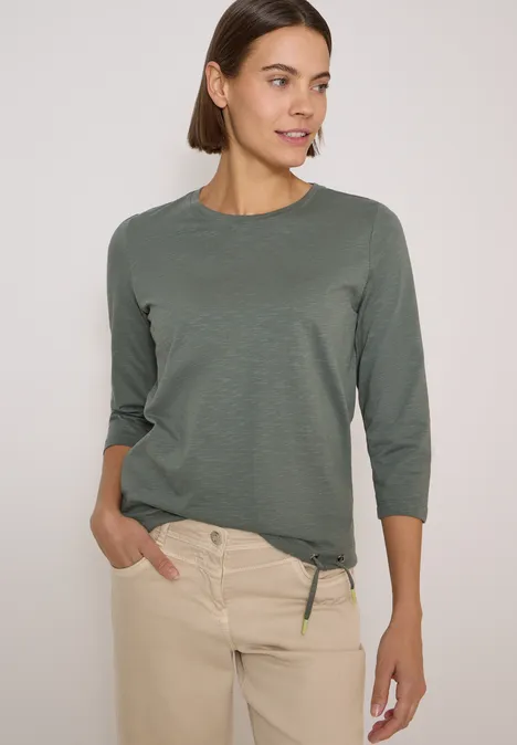 Frau in legerem olivgrünem 3/4-Ärmel-Shirt und beigen Hosen mit Händen in den Taschen.