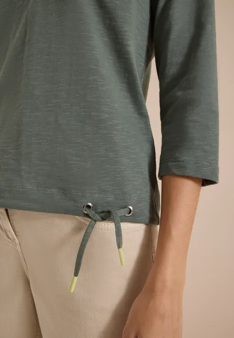 Grüner 3/4-Arm-Shirt mit Tunnelzug am Saum und neonfarbenen Kordelenden, kombiniert mit beigen Hosen.