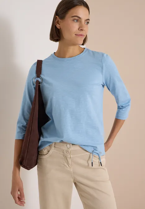 Frau im hellblauen 3/4-Arm-Shirt, beige Hose und brauner Umhängetasche, schaut seitlich vor hellem Hintergrund.