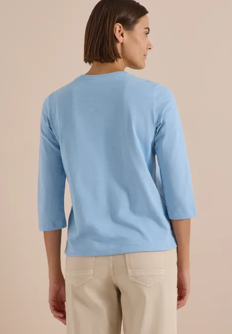 Rückansicht einer Frau in einem hellblauen Dreiviertelarm-Shirt und beigen Hosen vor beigem Hintergrund.
