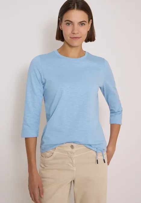 Frau trägt hellblaues Shirt mit 3/4-Ärmeln und beigefarbener Hose.