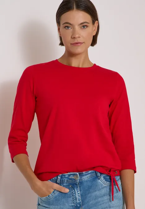 Frau vor hellem Hintergrund trägt roten Pullover mit 3/4-Ärmeln und blaue Jeans, eine Hand in Tasche.