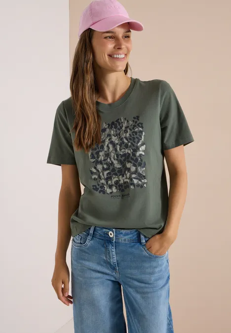 Kobieta uśmiechnięta w różowej czapce, oliwkowym t-shircie w kwiaty i niebieskich dżinsach.