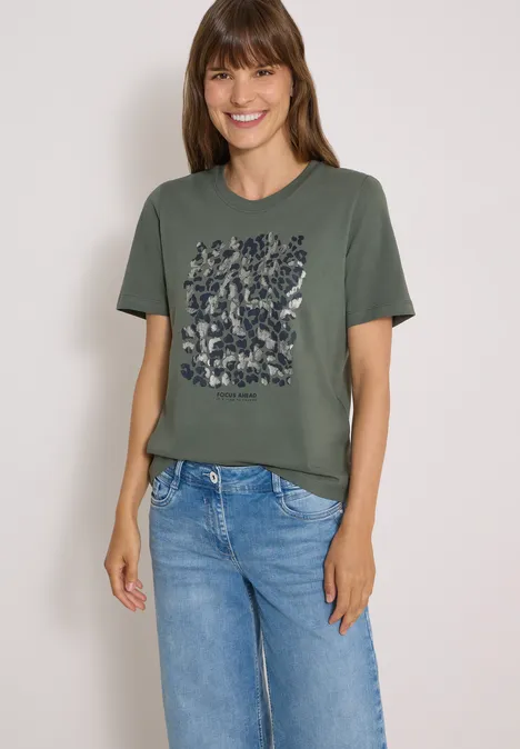 Kobieta uśmiechnięta w oliwkowym t-shircie z kwiatowym wzorem i niebieskich dżinsach na białym tle.