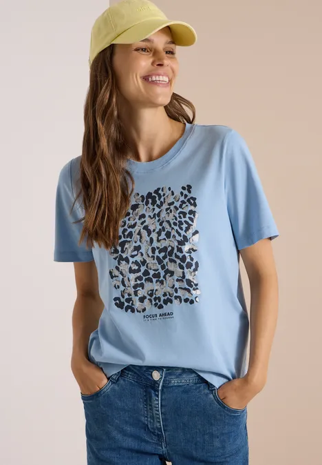 Uśmiechnięta kobieta w jasnoniebieskim t-shircie z panterkowym sercem, w dżinsach i żółtej czapce na neutralnym tle.