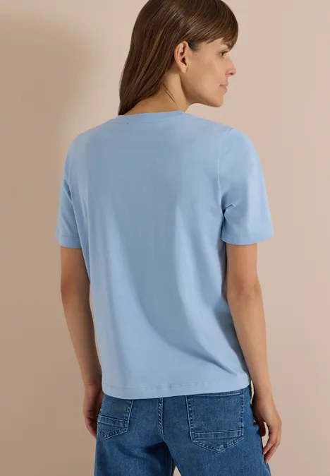 Kobieta widziana od tyłu w jasnoniebieskim T-shircie i dżinsach na neutralnym tle.